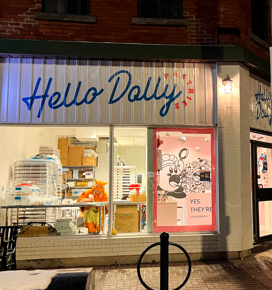 A photo of the Hello Dolly Pastries Storefront taken in the evening. The Shop is illuminated from within, and inside at the decorating table in front of the large window, the owner can be seen decorating cookies while wearing a Charmander onesie.
