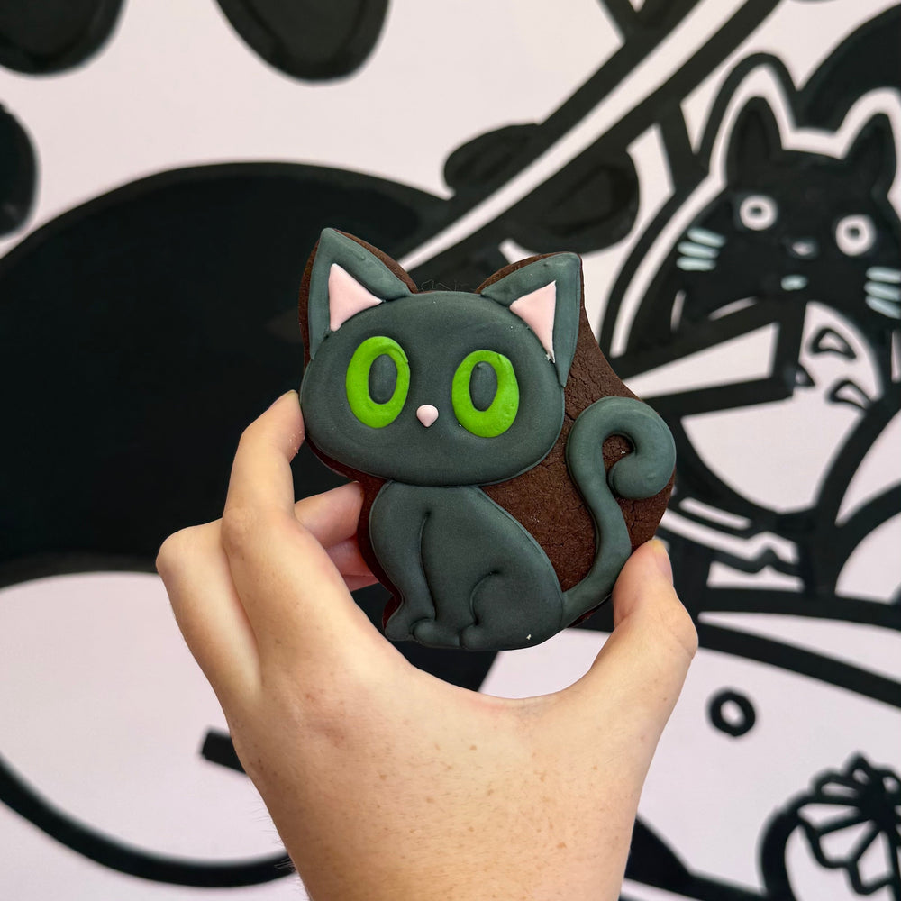 Hand holding a black cat-shaped cookie with green eyes against a black and white background with cat illustrations.