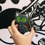 Hand holding a black cat-shaped cookie with green eyes against a black and white background with cat illustrations.