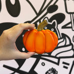 Hand holding a small orange pumpkin in front of a black and white Halloween-themed background